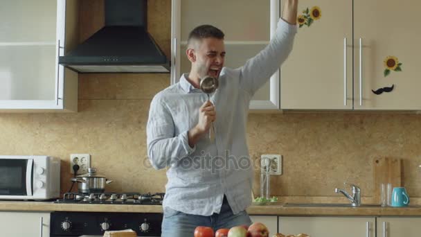 Slow motion of Attractive young funny man dancing and singing with ladle while cooking in the kitchen at home - Materiaali, video