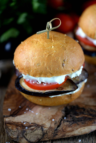 Hambúrgueres vegan com pão de milho com queijo macio, berinjela e tomates em um fundo de madeira
. - Foto, Imagem