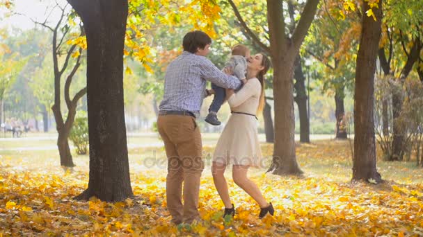 Αργή κίνηση χαρούμενος νέοι γονείς που αγκαλιάζει τους γιού στο Φθινοπωρινό πάρκο στο ηλιόλουστη μέρα - Πλάνα, βίντεο
