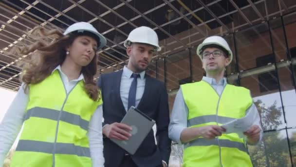 Team of Two Engineers and Businessman in Hard Hat Walking, Talking, and Using Tablet Computer. Glass Building or Skyscraper under Construction on Background. - Video, Çekim