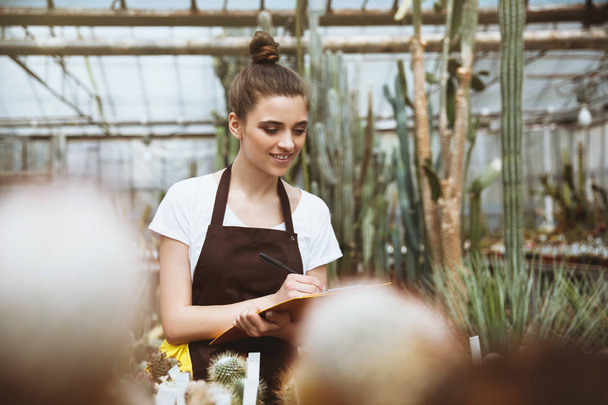 Happy young lady standing in greenhouse holding clipboard - Foto, afbeelding