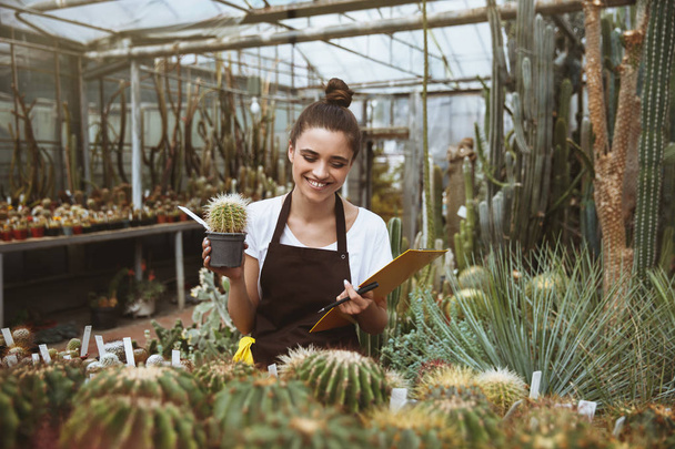 Happy young lady standing in greenhouse holding clipboard - Foto, Bild