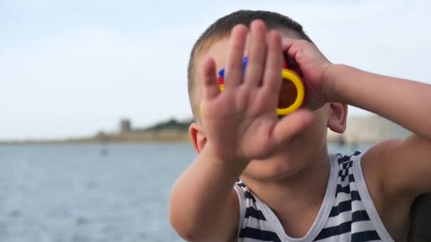 adorable kid w pasiastą koszulę patrząc przez lornetkę przed horyzont morze, macha ręką - Materiał filmowy, wideo