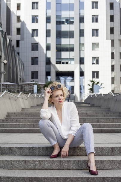 Beautiful blonde young woman wearing fashionable clothes sitting on stairs - Photo, image