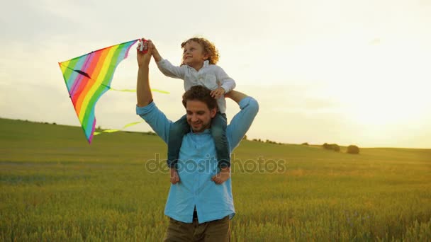 Feliz familia de padre joven caminando en el campo de trigo verde con su hijo en los hombros. Chico jugando con cometa voladora. Puesta de sol
 - Metraje, vídeo