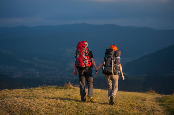Back view of couple hikers with backpacks holding hands and walk along a road with beautiful mountain landscape on background - Photo, Image