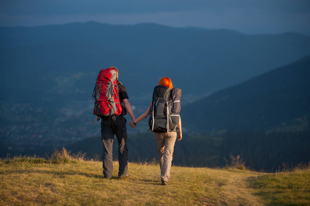 Rear view of couple hikers with backpacks holding hands and walking along a road with beautiful mountain landscape on background - Photo, Image