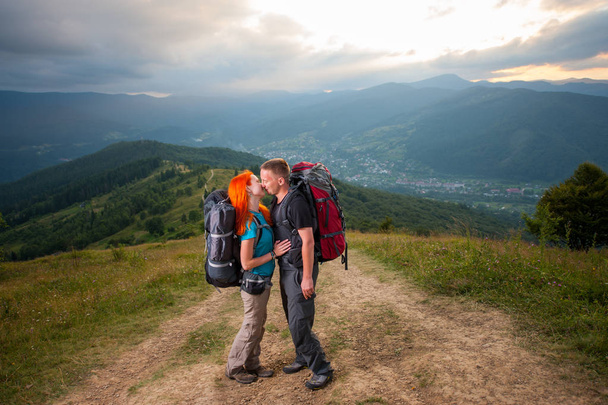 幸せなカップルのハイカーが山の道でキスします。背景の雄大な山々、森林、丘と雲の空に - 写真・画像
