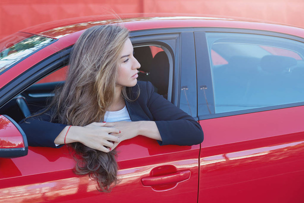 Portrait de jeune femme d'affaires caucasienne attrayante aux cheveux longs portant un costume formel au volant d'une voiture moderne rouge regardant de loin attendant que son patron vienne au bureau
. - Photo, image
