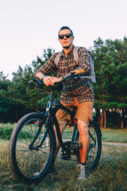 Красивый молодой человек велосипедист в солнцезащитных очках с велосипедом в летнем парке во время отпуска на закате
 - Фото, изображение