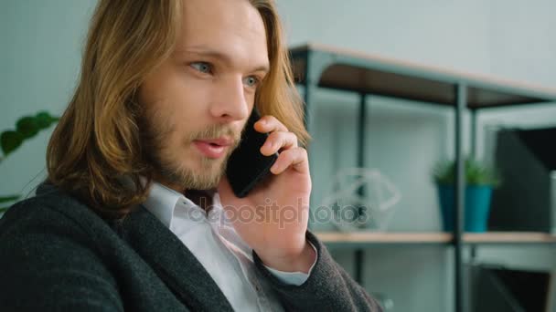 Young business man with long hair talking on a phone and working on a laptop computer. Close up - Кадры, видео