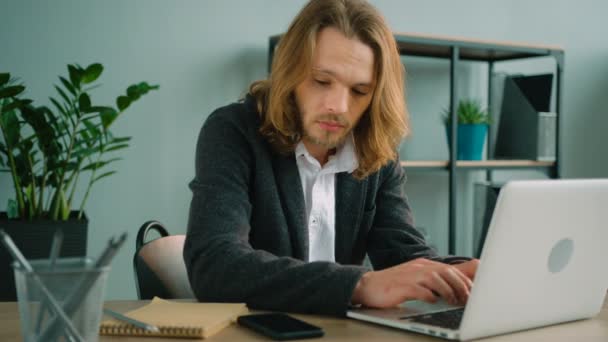 Mladý muž sedí v kanceláři, telefonování a psaní na přenosném počítači. Dlouhé vlasy muž dívá na obrazovku, Kontrola informací při rozhovoru na mobilní telefon. - Záběry, video