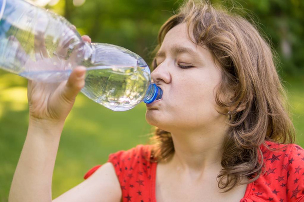 Νεαρή γυναίκα είναι πόσιμο νερό από το μπουκάλι στο ηλιόλουστο καυτή ημέρα. - Φωτογραφία, εικόνα