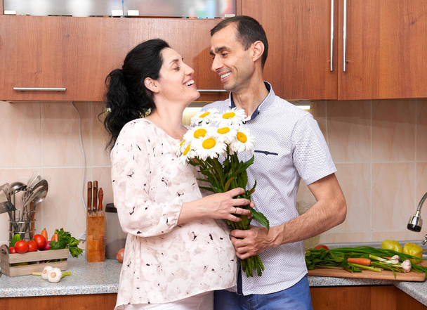 Hombre da flores de manzanilla a la mujer embarazada, pareja en el interior de la cocina con frutas y verduras frescas, concepto de alimentos saludables
 - Foto, imagen