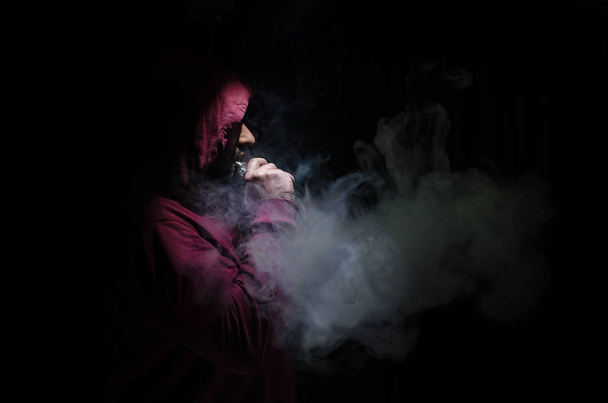 Vaping man holding a mod. A cloud of vapor. Black background. Vaping an electronic cigarette with a lot of smoke - Foto, afbeelding