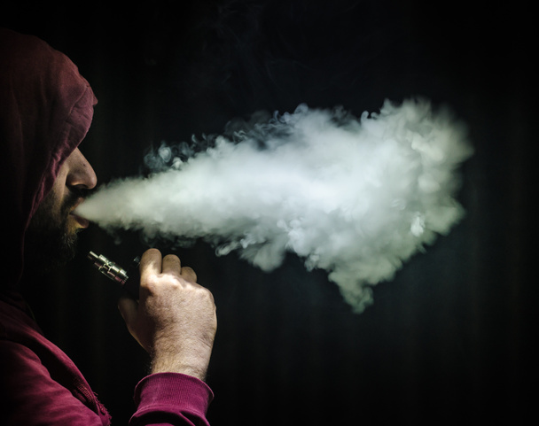 Hombre aspirante sosteniendo un mod. Una nube de vapor. Fondo negro. Vapeando un cigarrillo electrónico con mucho humo
 - Foto, imagen