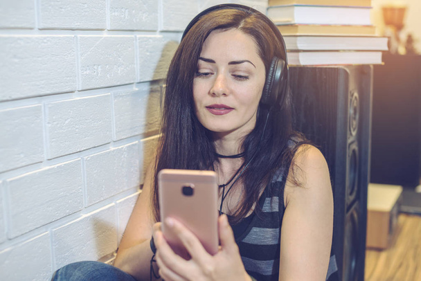 Woman słuchawki słuchanie audiobooka na telefon siedzi na tle ściany - Zdjęcie, obraz
