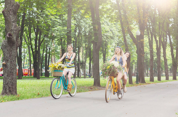 Happy boho chic girls ride together on bicycles in park - Φωτογραφία, εικόνα