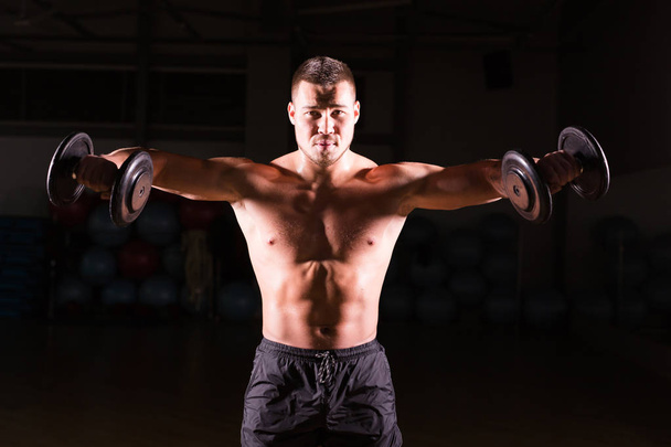 Handsome power athletic guy bodybuilder doing exercises with dumbbell. Fitness muscular body on dark background - Photo, Image