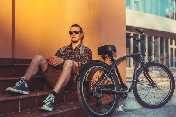 Όμορφος αρσενικό ποδηλάτη σε γυαλιά ηλίου με σακίδιο κάθεται σε βήματα κοντά το ποδήλατο ποδήλατο και ανάπαυσης Travel έννοια - Φωτογραφία, εικόνα