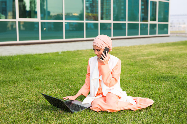 Uma jovem mulher de negócios muçulmana bonita em um parque enquanto conversa no smartphone olhando para o laptop
 - Foto, Imagem