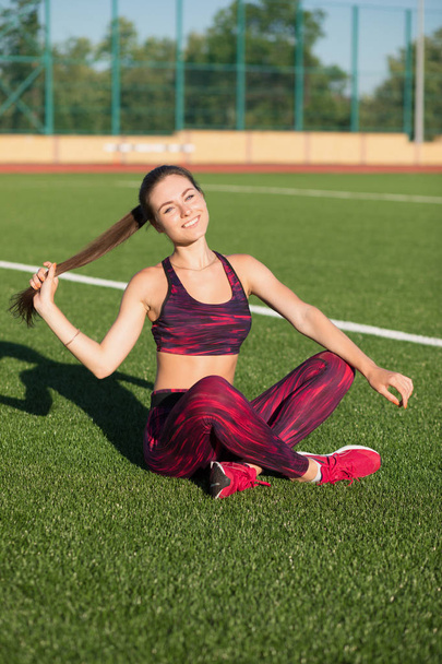 Young smiling sportswoman in sportswear sitting on stadium green grass outdoors. Healthy lifestyle concept. Summer sport activity. - Foto, afbeelding