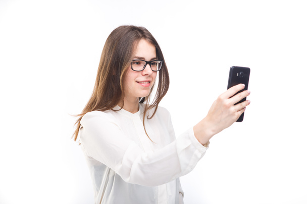 Mooi meisje in glazen en een wit overhemd neemt een foto van zichzelf op de cameratelefoon, selfie op witte geïsoleerde achtergrond - Foto, afbeelding
