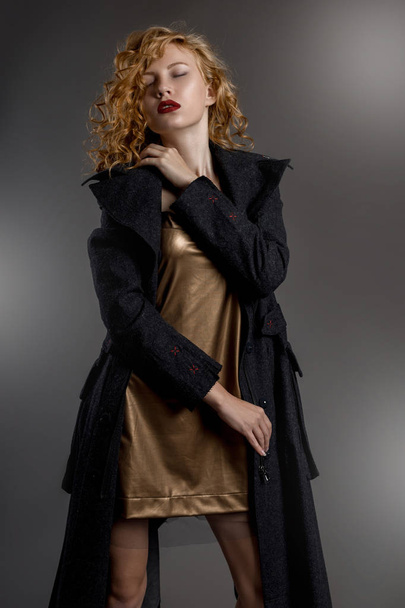 Sexy chica de piernas largas con hermoso cabello y hermosos ojos en un seductor vestido corto de oro y abrigo gris en estilo gótico
 - Foto, imagen