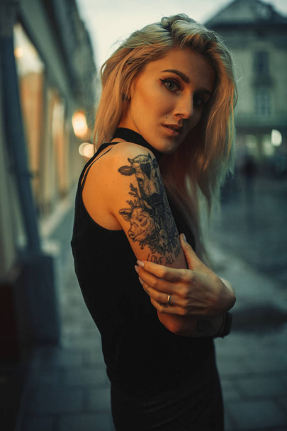 Портрет молодой женщины с татуировкой на плече, стоящей на городской улице вечером
. - Фото, изображение