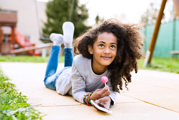 Beautiful african american girl with smart phone, eating lollipo - Foto, Imagen