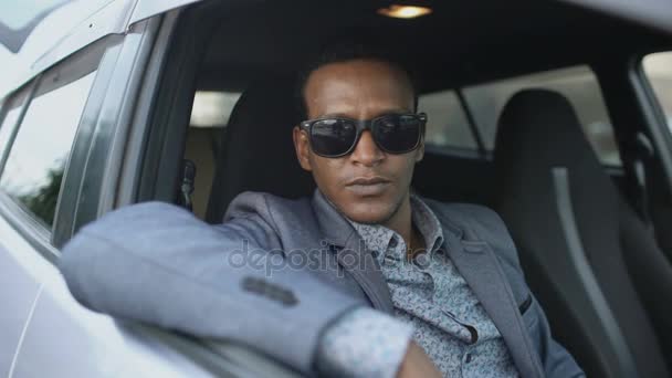 Portrait of serious businessman in sunglasses sitting inside car and looking into camera outdoors - Materiaali, video