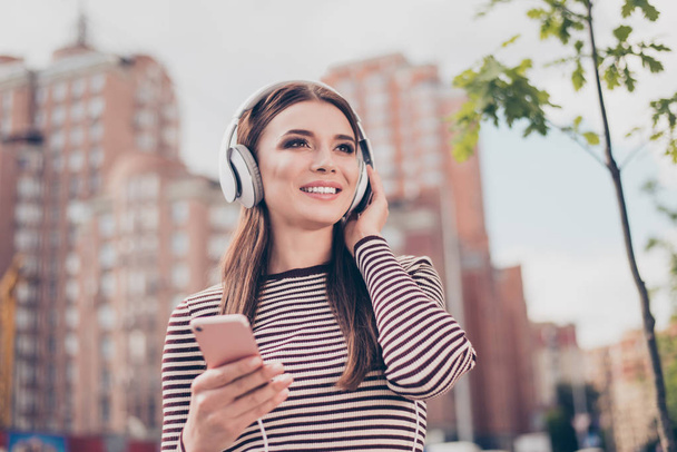 Carefree young lady is listening to favourite song on her smartphone outdoors, walking on the spring street, wearing cozy outfit, modern headphones - Zdjęcie, obraz