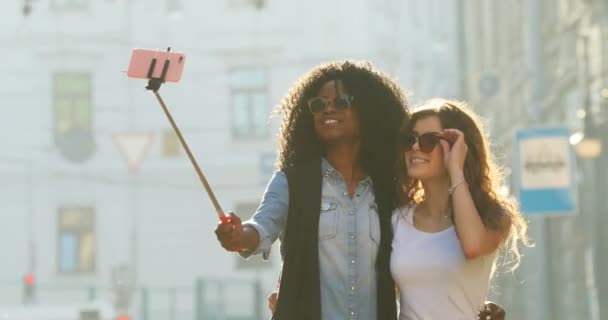 Nahaufnahme Porträt von zwei schönen lächelnden Freundinnen, die eine Sonnenbrille korrigieren und das Zeichen Frieden beim Fotografieren mit dem Selfie-Stick während des Spaziergangs zeigen. - Filmmaterial, Video