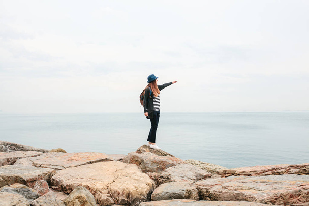 Girl traveler in a hat with a backpack next to the sea showing a hand into the distance. Travel, leisure, hiking. - Photo, Image