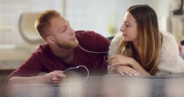 Young cheerful couple lying on sofa listening music on smartphone - Video