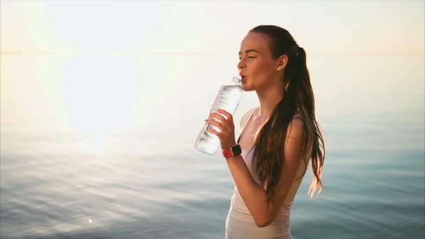 Jovem mulher esportiva bebendo uma água da garrafa de plástico perto do mar
. - Filmagem, Vídeo