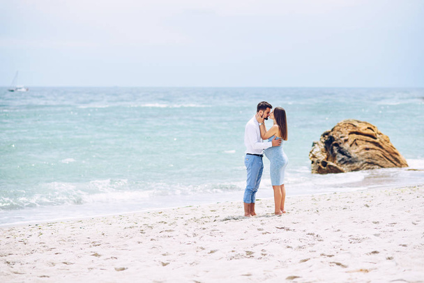 A beautiful pregnant woman in a blue dress kissing to the nose her courageous and handsome husband in a shirt and blue trousers against the background of the rocks and the sea. - Фото, изображение
