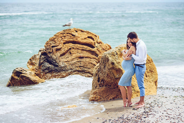 A beautiful pregnant woman in a blue dress and her courageous and handsome husband in a shirt and blue trousers leaned to each other against the background of the rock with seagull and the sea. - Foto, imagen