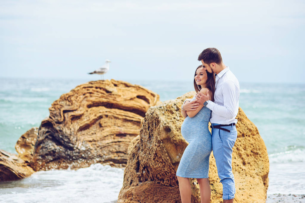 A beautiful pregnant woman in a blue dress and her courageous and handsome husband in a shirt and blue trousers leaned to each other against the background of the rocks with seagull and the sea. - Фото, зображення