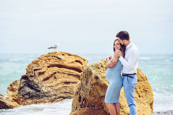 A beautiful pregnant woman in a blue dress and her courageous and handsome husband in a shirt and blue trousers leaned to each other against the background of the rock with seagull and the sea. - Φωτογραφία, εικόνα