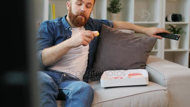Hipster man watching tv and eating a lot of doughnuts while relaxing on sofa at home in the living room. - Video