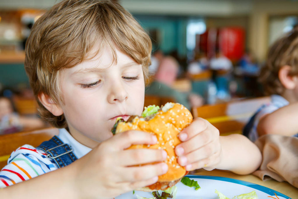 Χαριτωμένο υγιή προσχολικής ηλικίας αγόρι τρώει χάμπουργκερ κάθεται σε σχολικό κυλικείο - Φωτογραφία, εικόνα