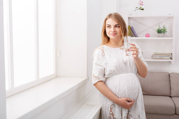 Беременная женщина со стаканом воды сидит на диване
 - Фото, изображение