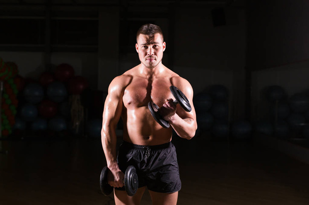 Jóképű atlétikus srác testépítő gyakorlatokat végez súlyzóval. Fitness izmos test sötét háttér - Fotó, kép