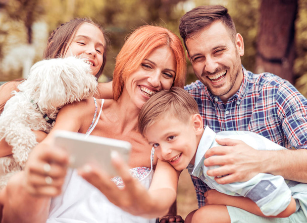 Szczęśliwa rodzina z psem biorąc selfie przez inteligentny telefon w parku - Zdjęcie, obraz