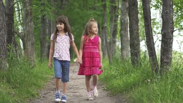 two little girls hold hands and walk down the green alley. children walk outdoors. two little sisters. slow motion - Footage, Video