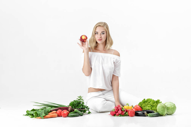 Beautiful blond woman in white clothes and lots of fresh vegetables on white background. girl is eating nectarine - Fotó, kép