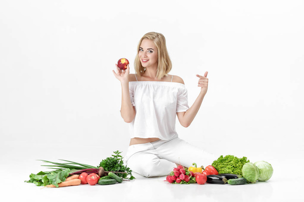 Beautiful blond woman in white clothes and lots of fresh vegetables on white background. girl is eating nectarine - Foto, Bild