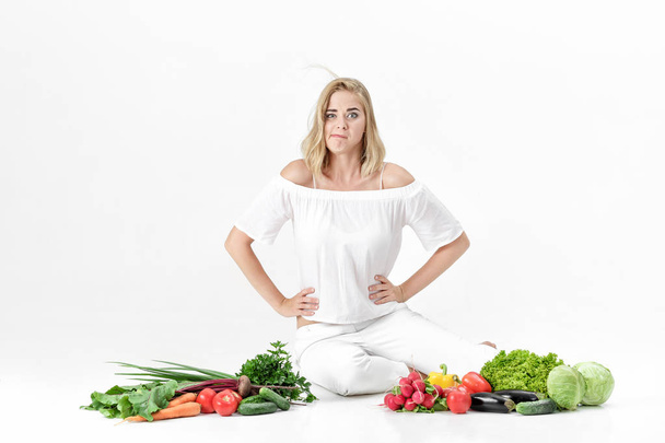 Beautiful upset blond woman in white clothes and lots of fresh vegetables on white background. diet - Fotó, kép