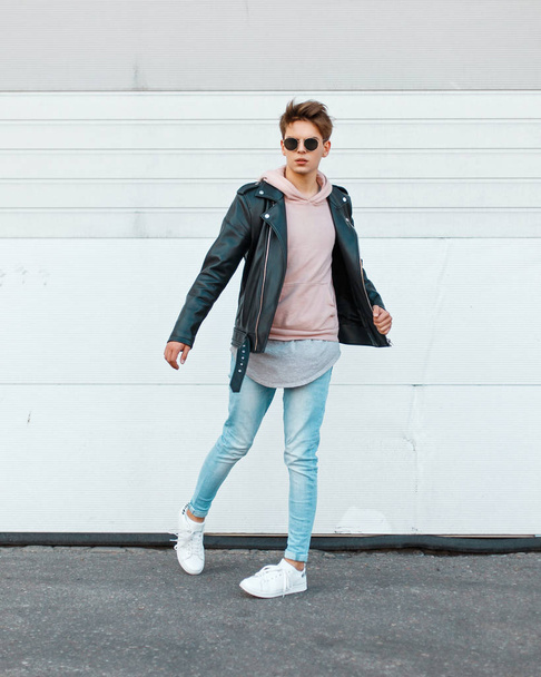 Young guy in a stylish black leather jacket, a pink sweatshirt, blue gin and white shoes in motion - 写真・画像